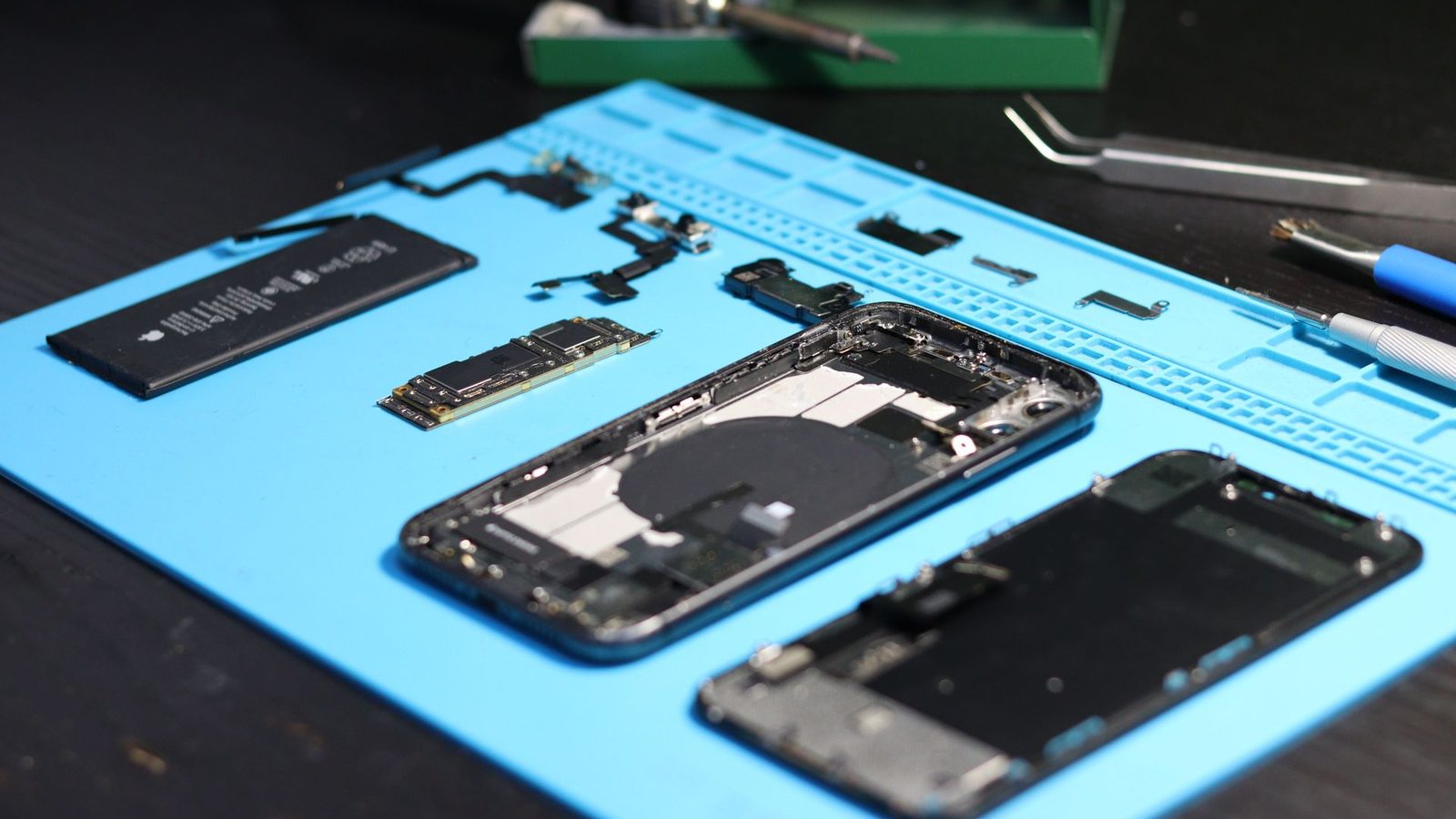 An iPhone stripped down on a repair mat at iComm Solutions — battery, logic board, flex cables and the screen assembly laid out beside the chassis, with tweezers and a spudger to hand
